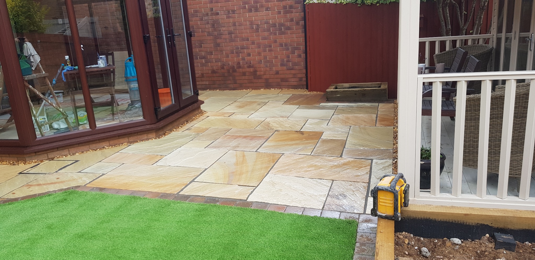 Ułożona z piaskowca patio z obrzeżem z cegieł, widok na zewnątrz z widocznymi narzędziami pracy i sztuczną trawą.