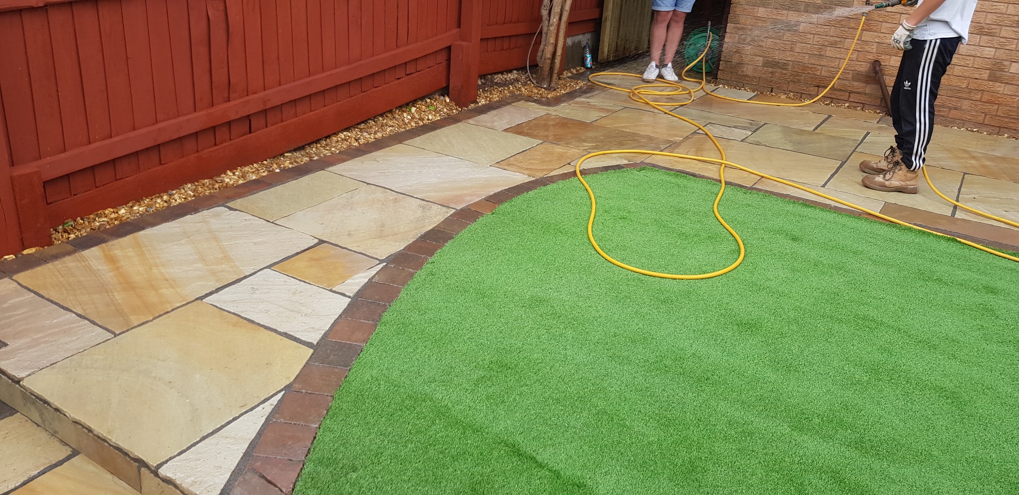 Umyta nawierzchnia z piaskowca i sztucznej trawy w ogrodzie, żółty wąż ogrodowy zwinięty w zygzak, brązowe obrzeża z kostki, czerwone drewniane ogrodzenie w tle.