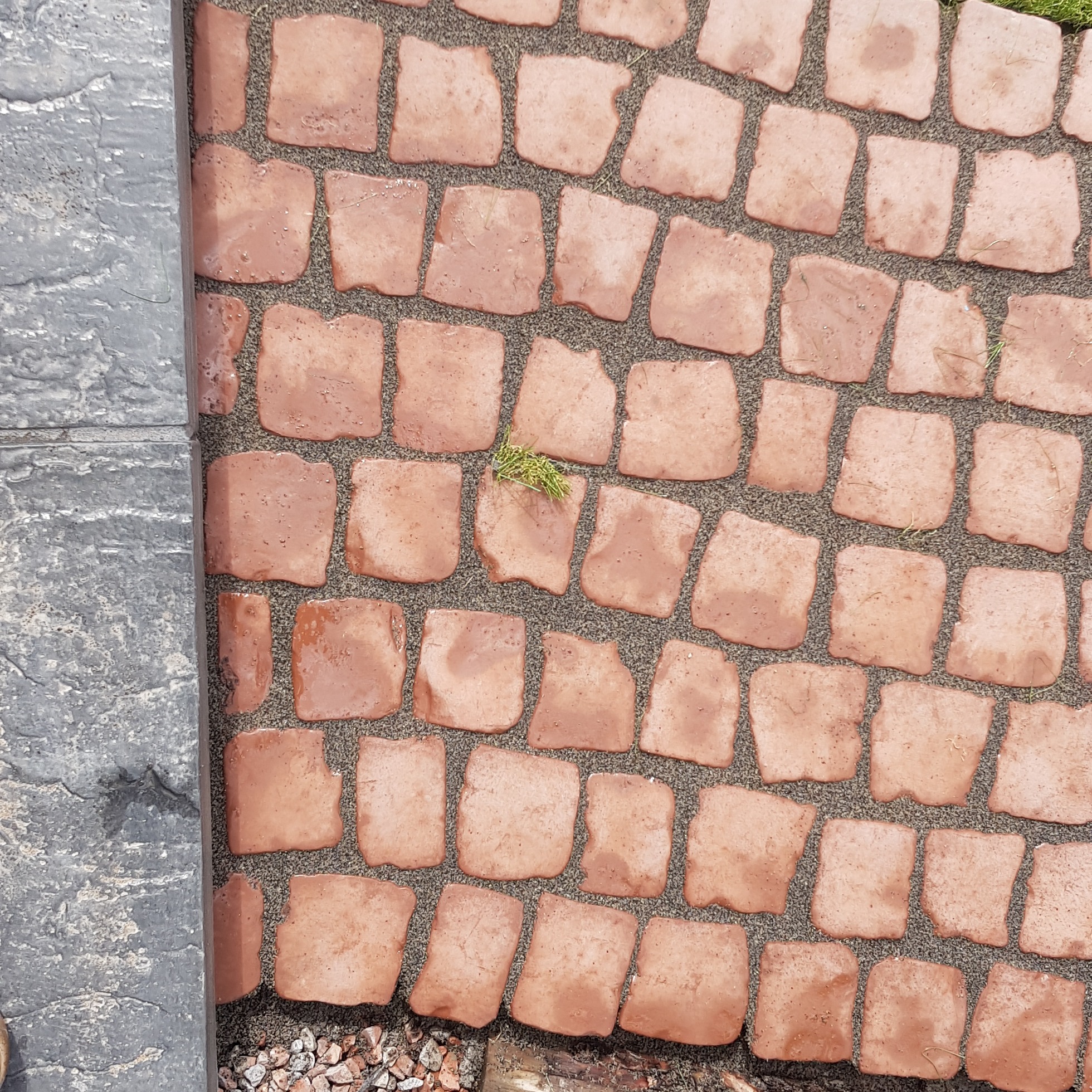 Fragment nawierzchni z nieregularnych, czerwono-brązowych kostek ułożonych na szarym podłożu, z fragmentem szarej, betonowej obwódki po lewej stronie i drobnymi kamykami w rogu.