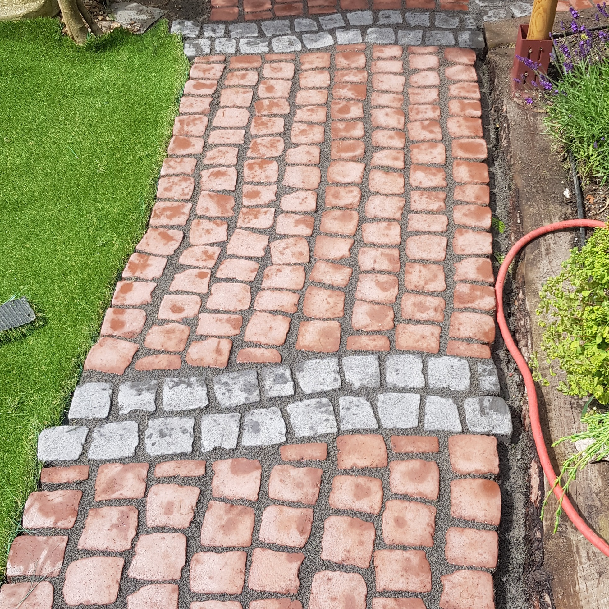 Ścieżka z czerwonej kostki brukowej oddzielona szarymi obrzeżami, wypełniona piaskiem, przylegająca do trawnika i rabaty z roślinami.