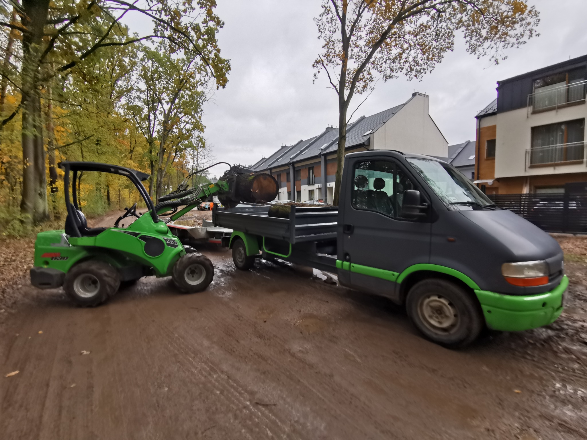 Zielony miniładowarka z chwytakiem transportuje pień drzewa na przyczepę ciężarówki na błotnistej drodze, w tle zabudowa szeregowa i drzewa.