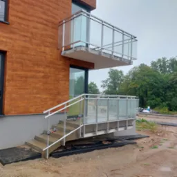 Nowoczesny budynek z drewnianą elewacją i balkonem z balustradą szklaną oraz schodami z poręczami, widoczne przyssawki do szkła na poręczy schodowej, teren wokół w trakcie prac wykończeniowych.