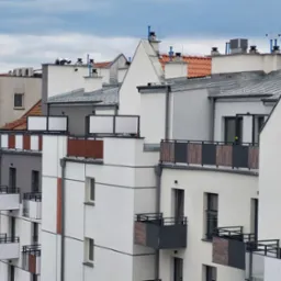 Widok na nowoczesne budynki mieszkalne z balkonami o różnych wykończeniach, z dominującą białą elewacją i elementami szarości oraz drewna, na tle pochmurnego nieba.