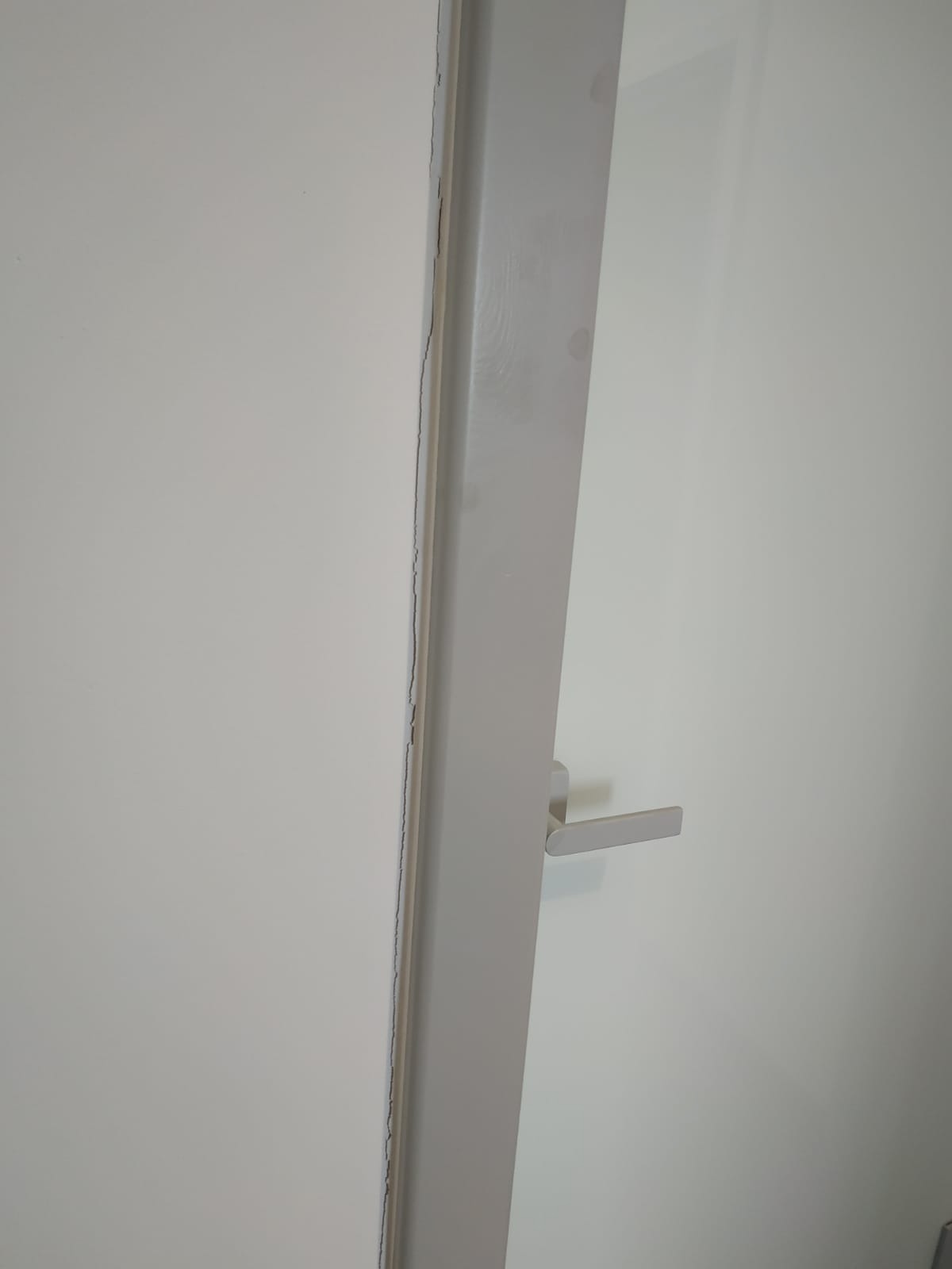 Szczegółowe ujęcie białych drzwi z widocznym pęknięciem na ościeżnicy, z klamką w kolorze białym i fragmentem białej ściany w tle.