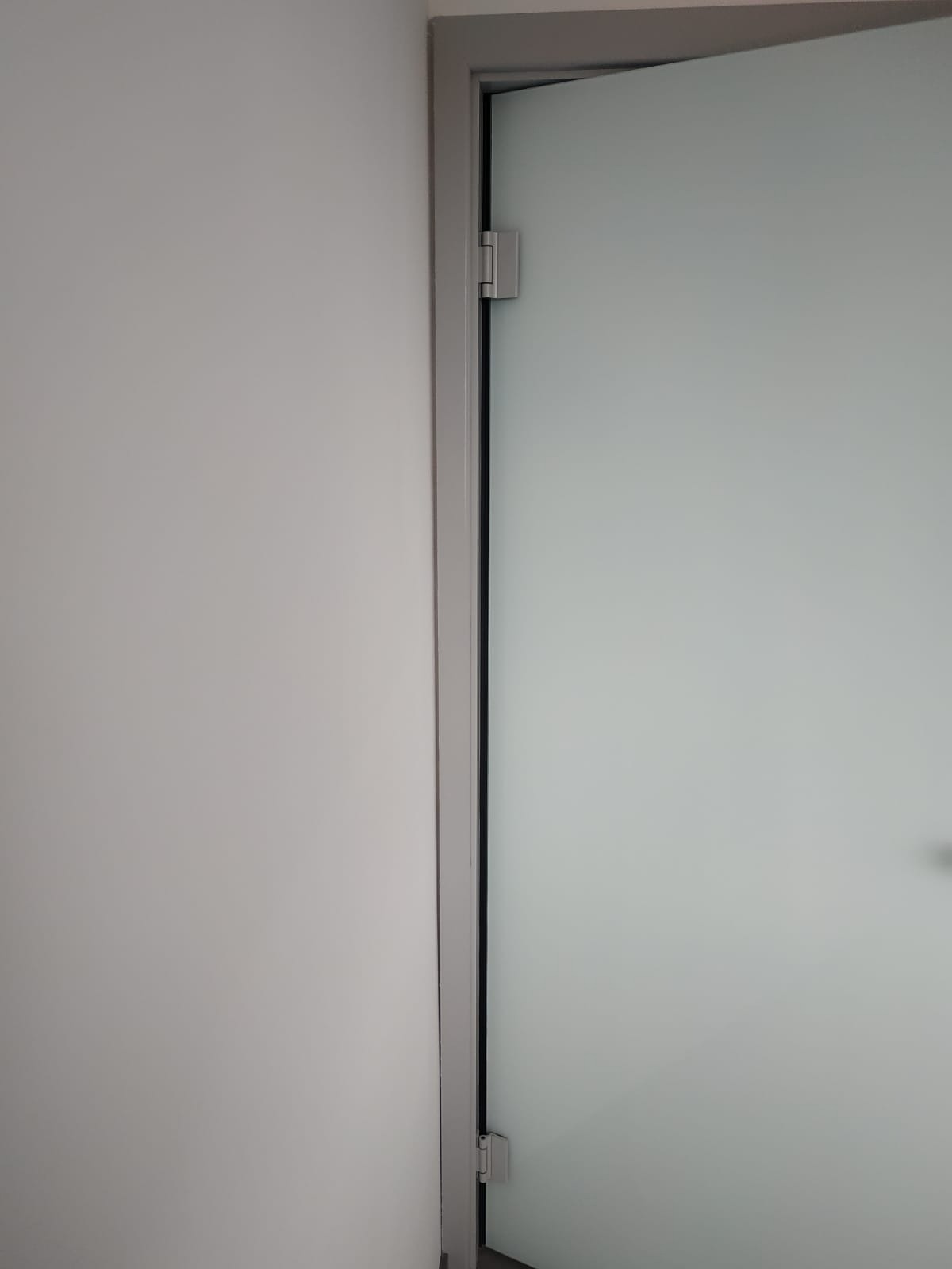 Detal wewnętrzny: częściowo otwarta, matowa, szklana tafla w szarej ramie, zawieszona na zawiasach, przylegająca do jasnoszarej ściany.