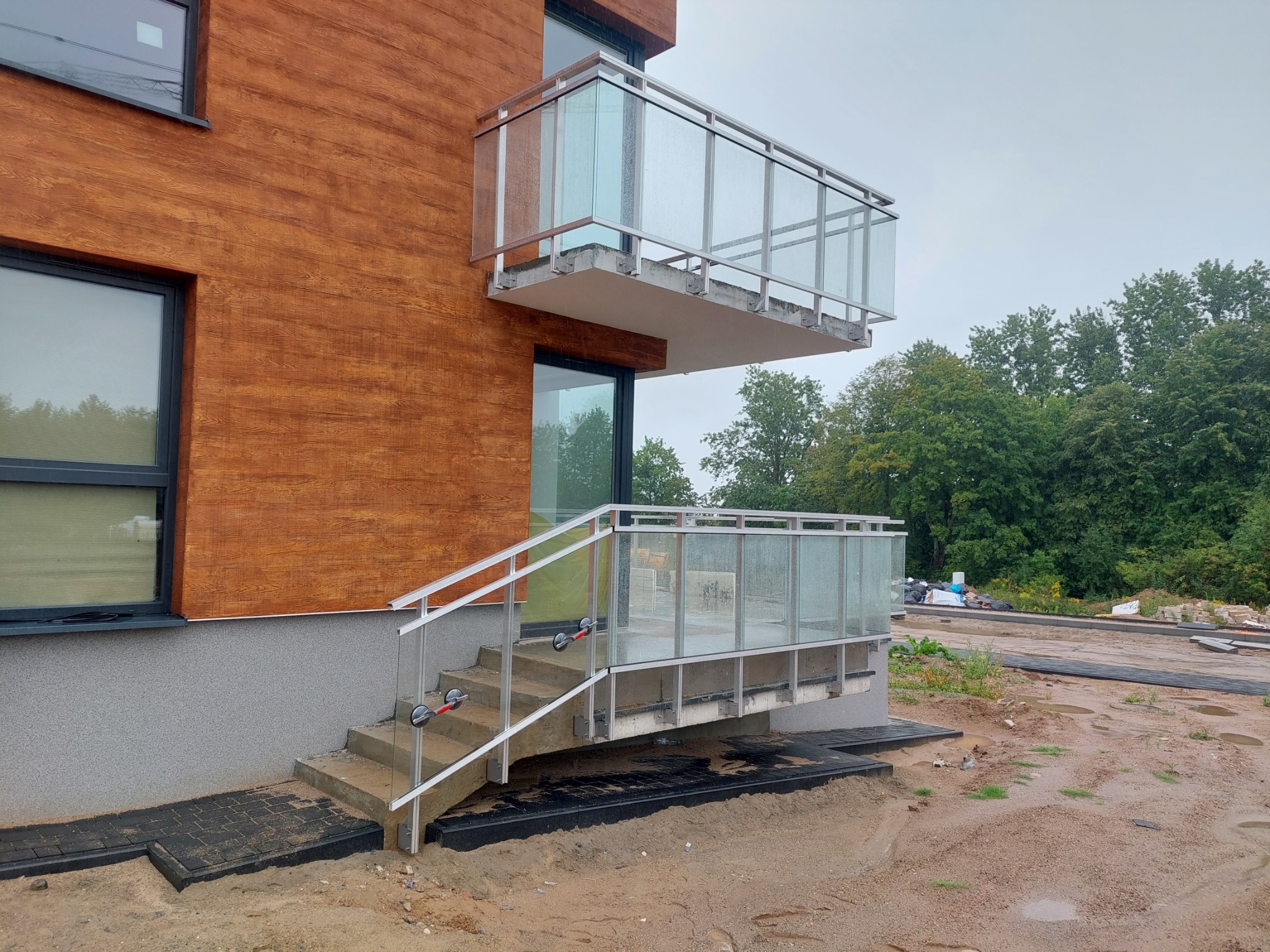 Nowoczesny budynek z drewnianą elewacją i balkonem z balustradą szklaną oraz schodami z poręczami, widoczne przyssawki do szkła na poręczy schodowej, teren wokół w trakcie prac wykończeniowych.