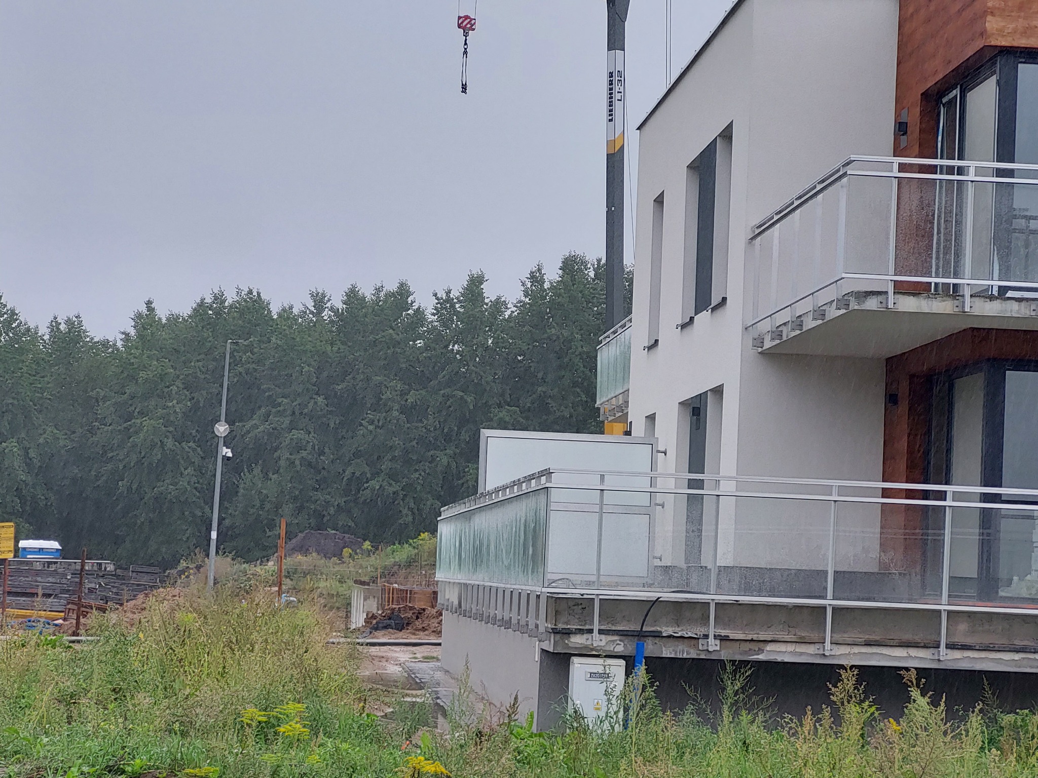 Nowoczesny budynek z balkonami zabezpieczonymi szklanymi panelami i metalowymi poręczami, widok z poziomu gruntu na tle drzew i placu budowy z żurawiem.