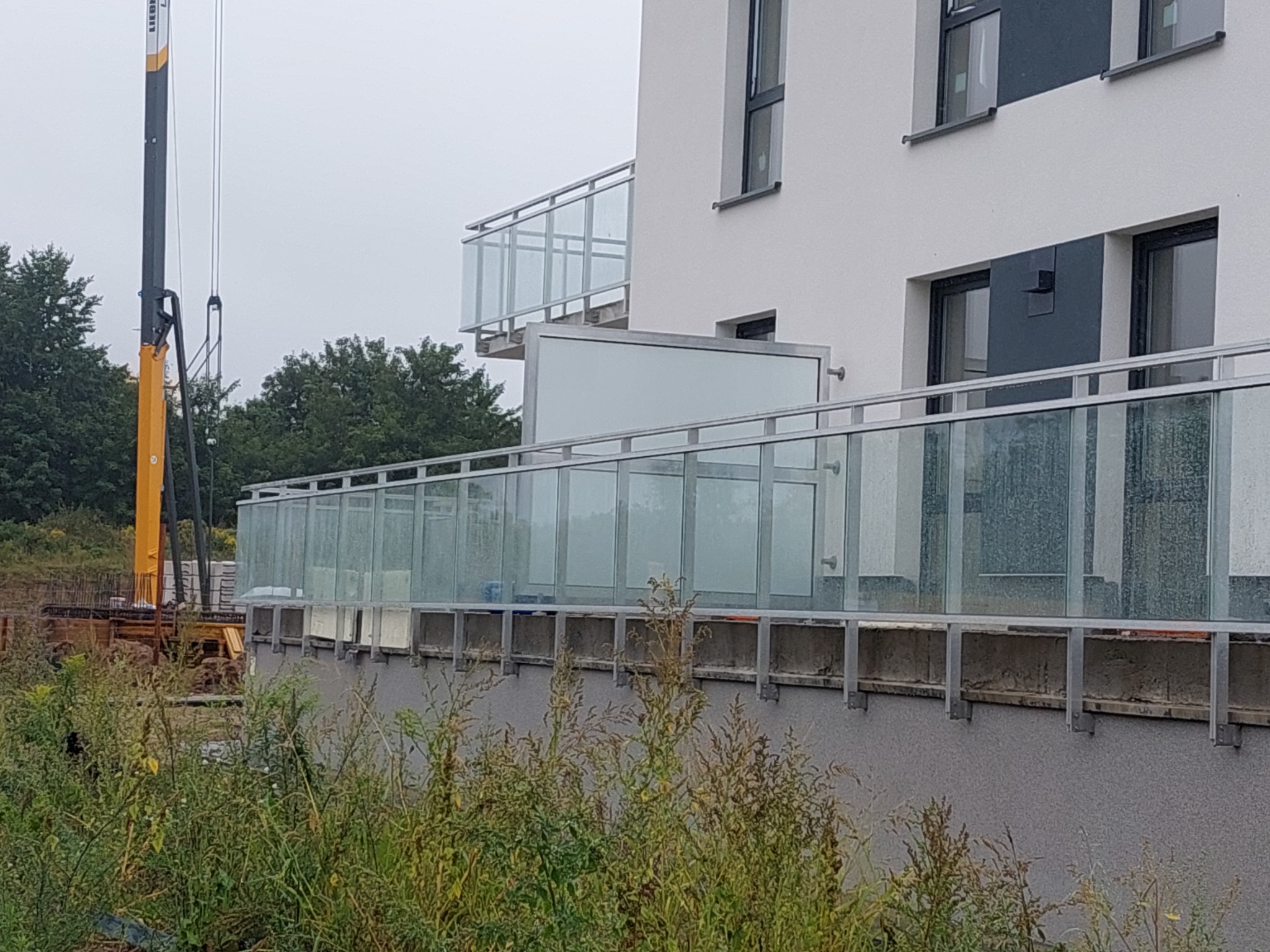 Szklane balustrady balkonowe z metalowymi poręczami w nowym budynku, widok z dołu na tle budowy z żurawiem, elementy montażowe widoczne.