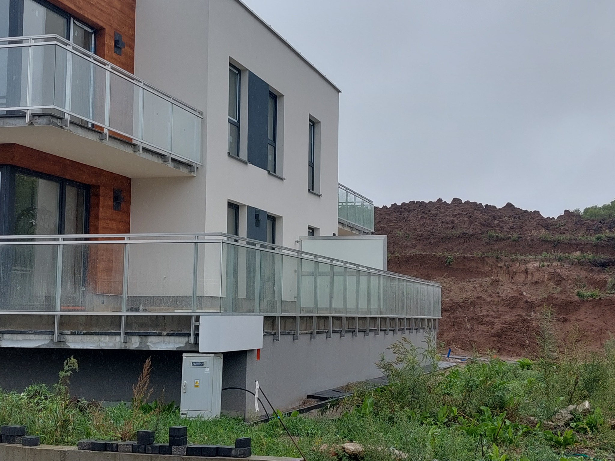 Nowoczesny budynek z balkonami z balustradami szklanymi, widoczne detale mocowań i konstrukcji, otoczenie w stanie surowym z ziemią i zielenią.