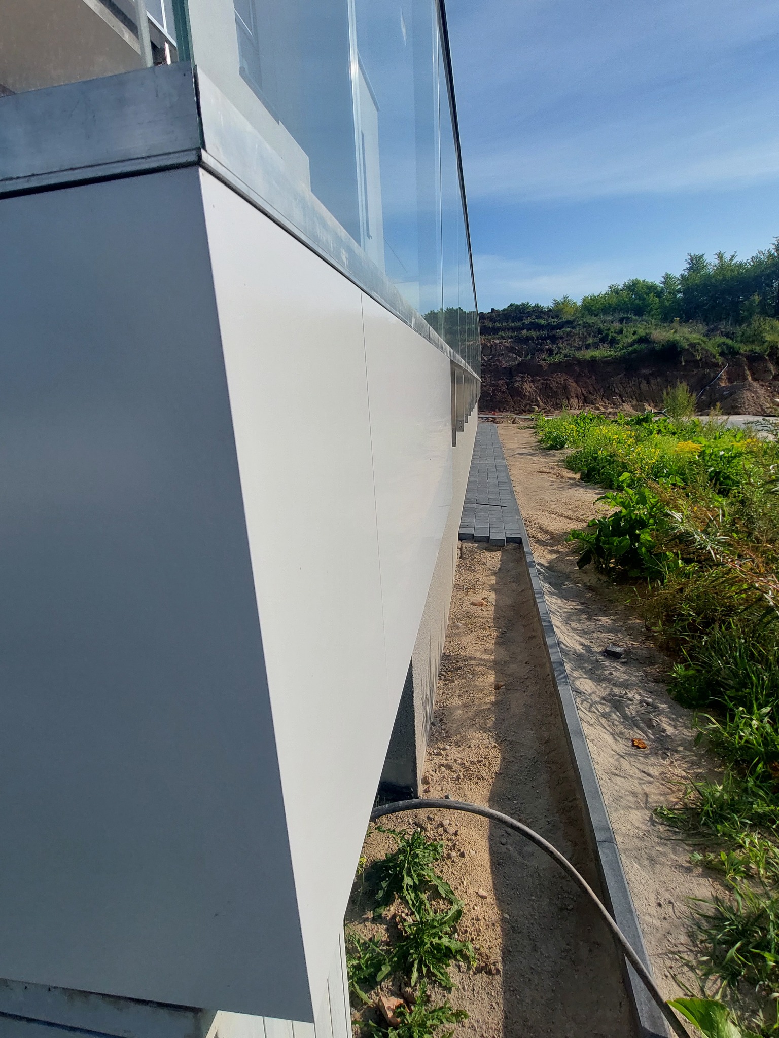 Szklana balustrada balkonowa z widocznymi elementami mocującymi, zewnętrzna strona budynku z zagospodarowaniem terenu, w tle skarpa z roślinnością.