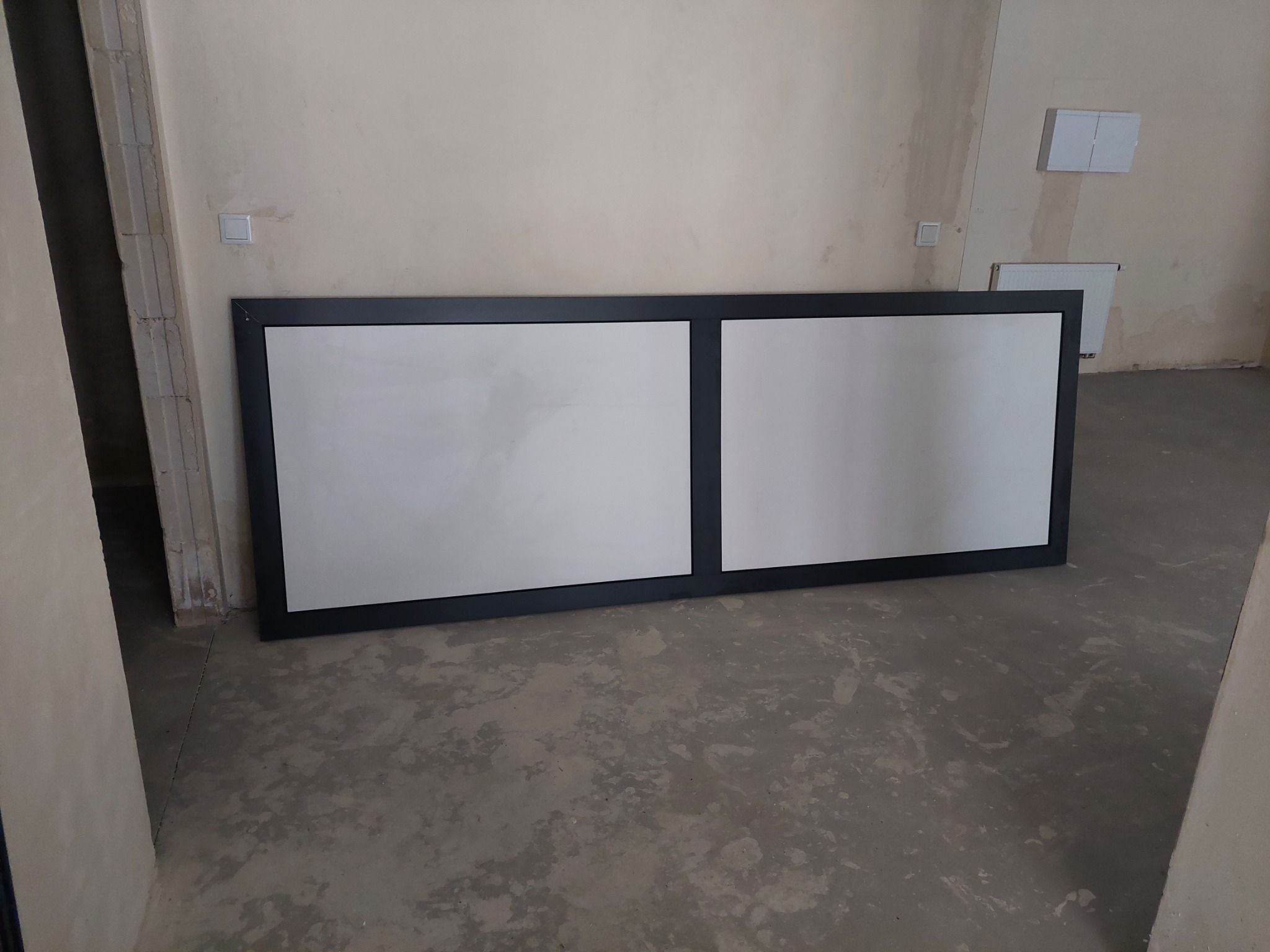 Dwa panele szklane w czarnej ramie, ustawione na podłodze w surowym wnętrzu, widoczne ślady po remoncie na ścianach i podłodze.