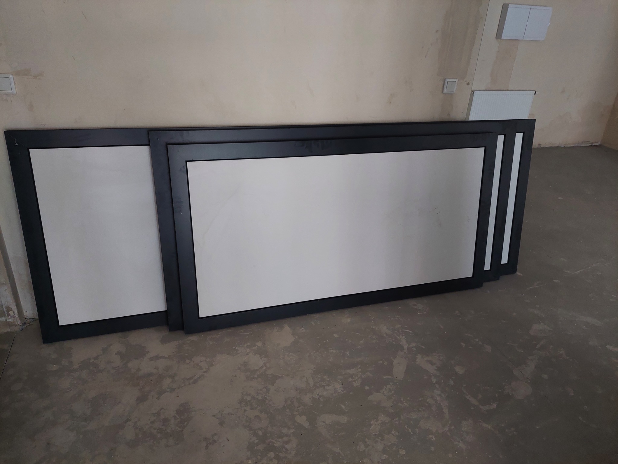 Trzy prostokątne konstrukcje z ciemną ramą i białym wypełnieniem, opierające się o ścianę w surowym wnętrzu, prawdopodobnie przygotowane do montażu.