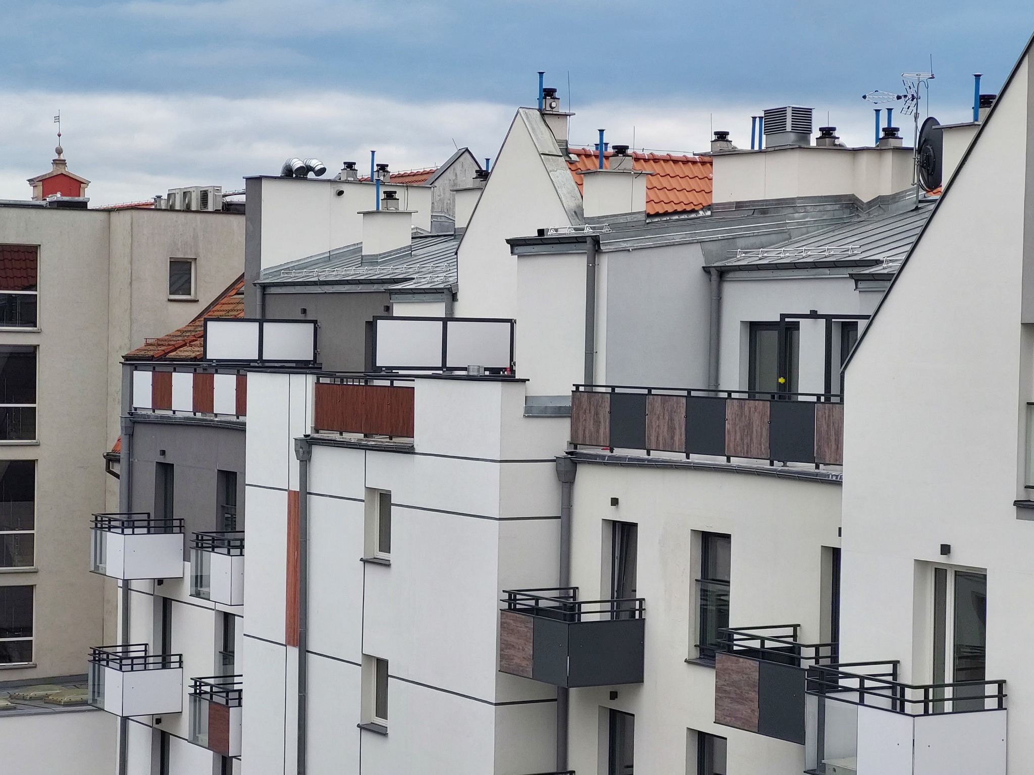 Widok na nowoczesne budynki mieszkalne z balkonami o różnych wykończeniach, z dominującą białą elewacją i elementami szarości oraz drewna, na tle pochmurnego nieba.