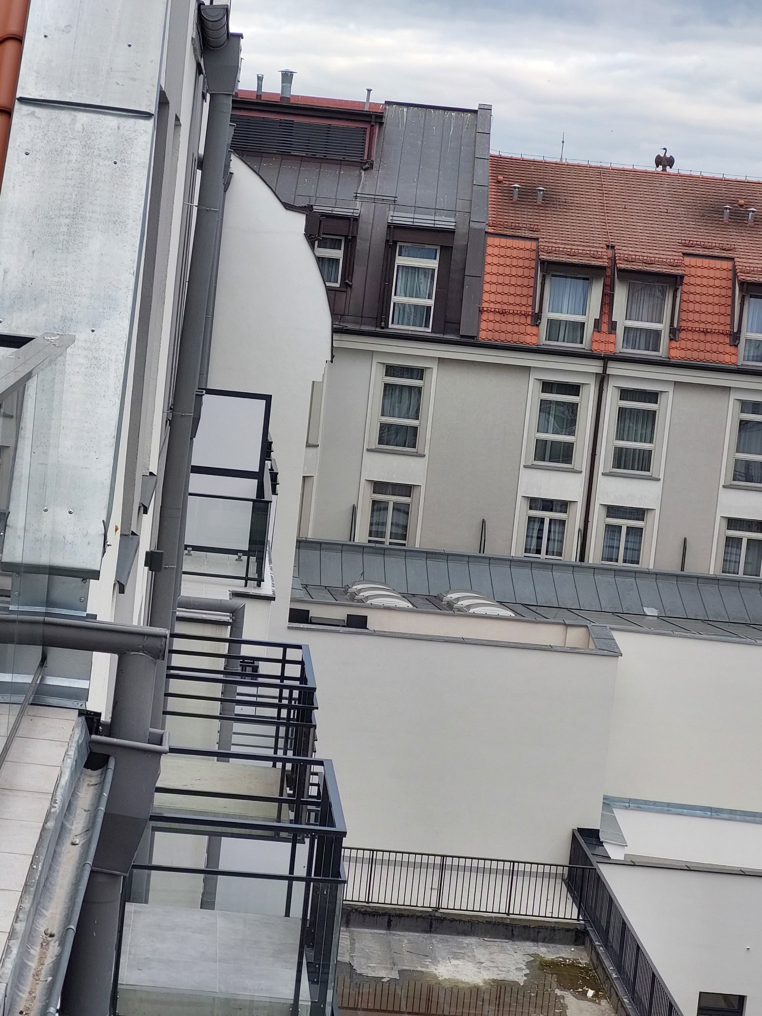 Widok z góry na nowoczesne balkony z czarnymi, metalowymi barierkami, z fragmentami budynków w tle, w tym jeden z czerwoną dachówką i figurą ptaka na szczycie.