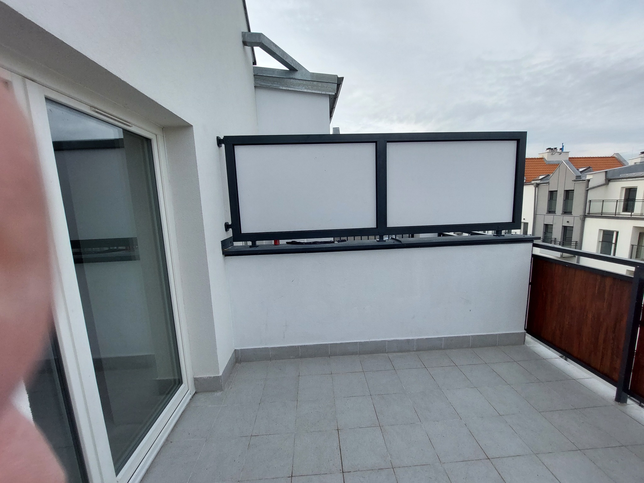 Balkon z nowoczesną balustradą z wypełnieniem z białych paneli w czarnej ramie oraz drewnianymi elementami, widok na budynki mieszkalne w tle, szare płytki na podłodze.