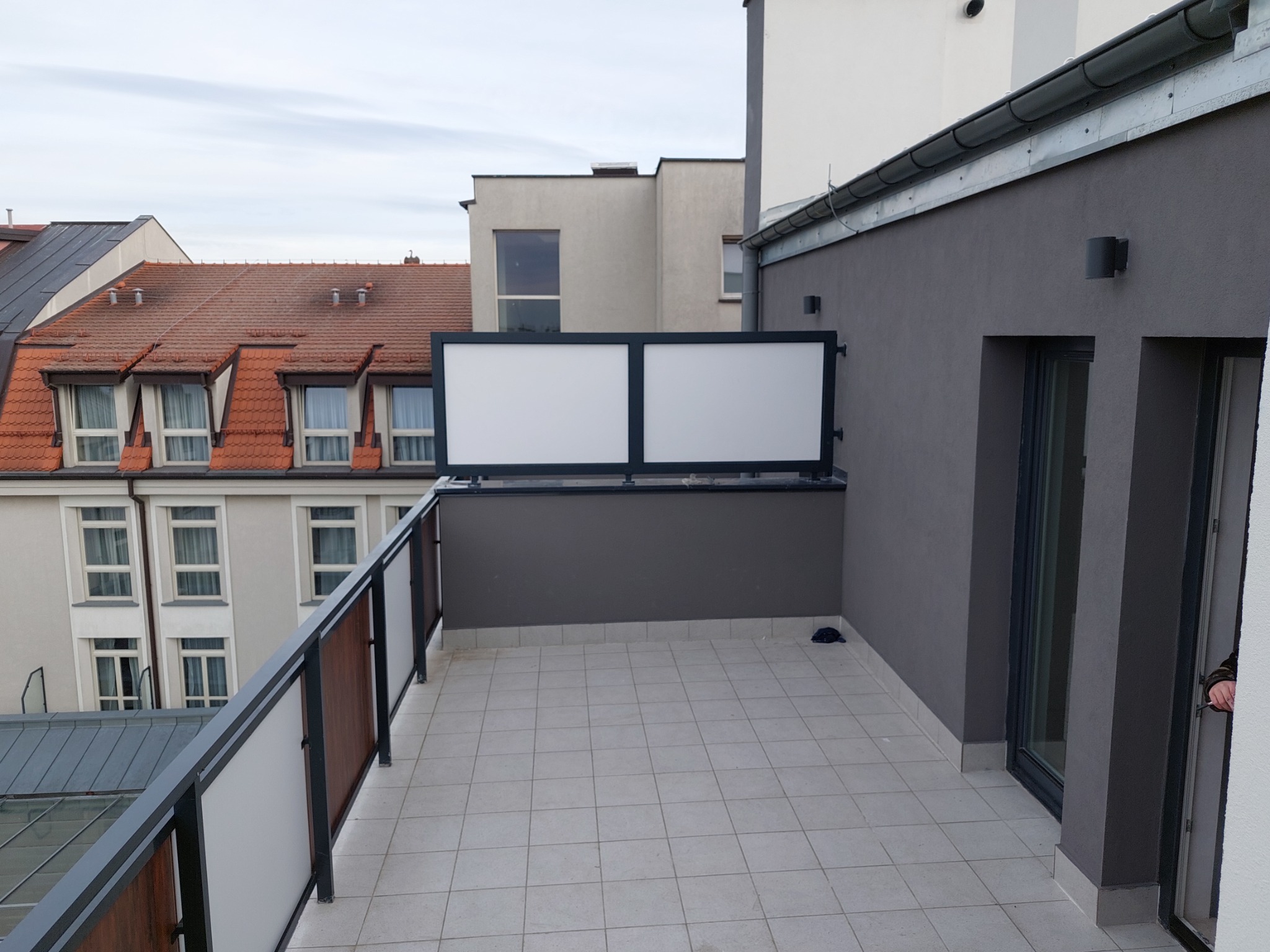 Nowoczesny balkon z szarą balustradą z elementami imitującymi drewno i białymi panelami, widok na sąsiednie budynki z czerwonymi dachami.