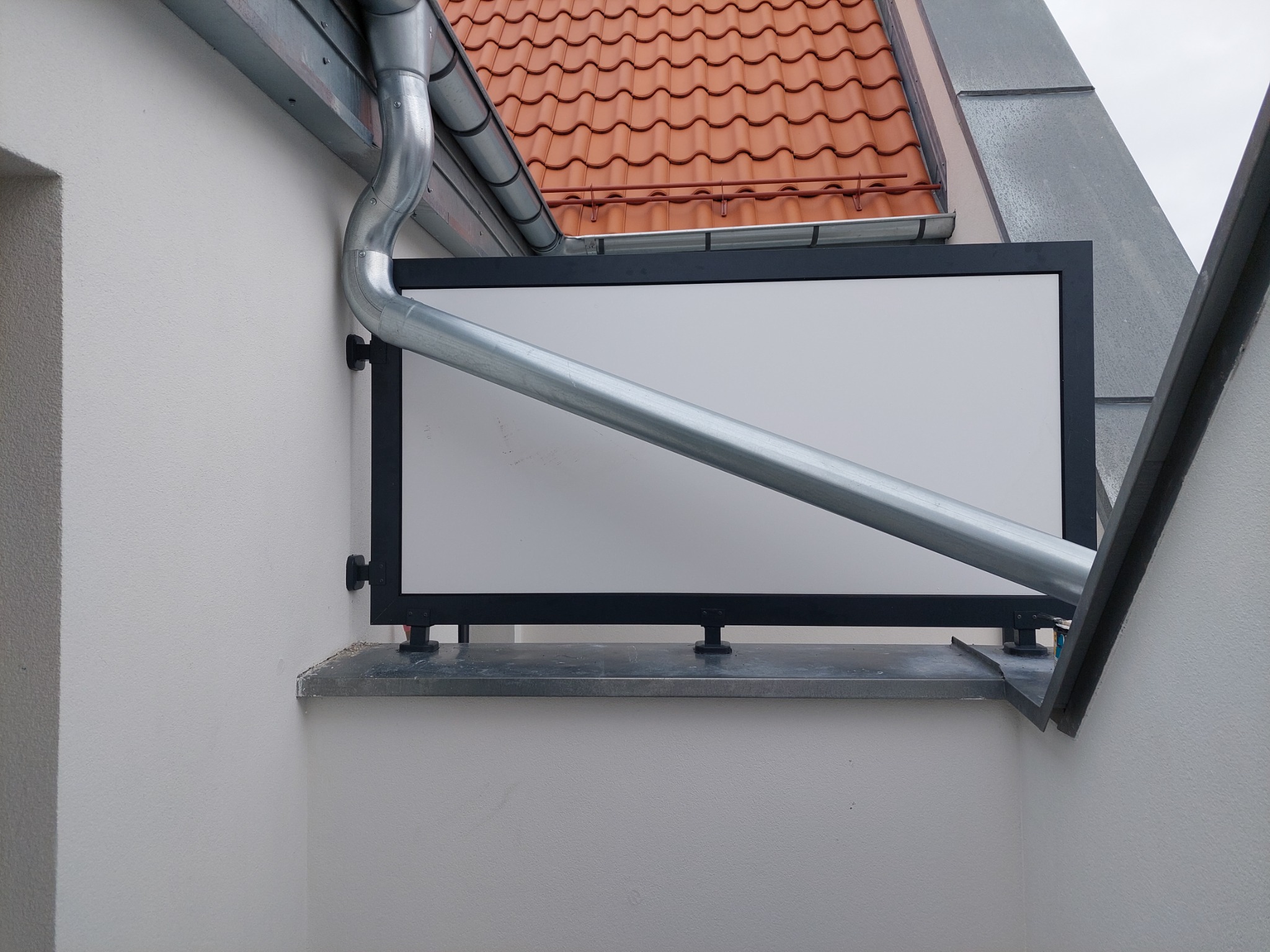 Balkon z czarną ramą i białym wypełnieniem, widok z dołu, rynna przechodząca ukośnie na tle, dach z czerwonej dachówki w tle.