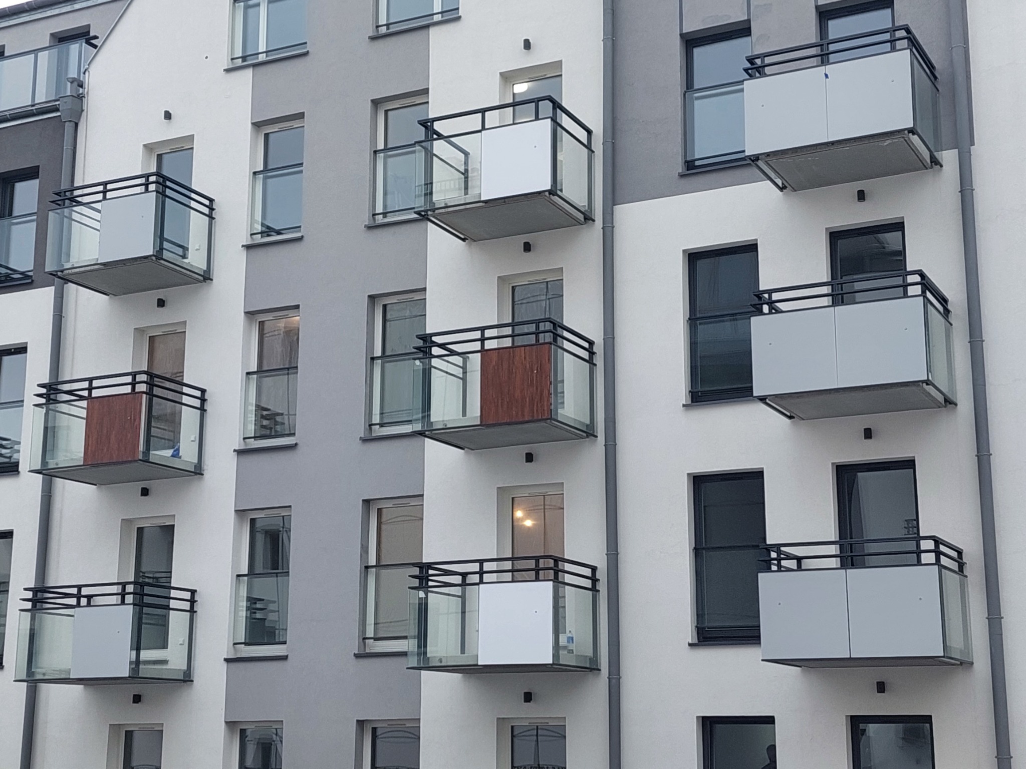 Fasada budynku wielorodzinnego z balkonami o zróżnicowanym wykończeniu: balustrady szklane i z płyt drewnopodobnych, czarne metalowe ramy, biało-szara kolorystyka elewacji.