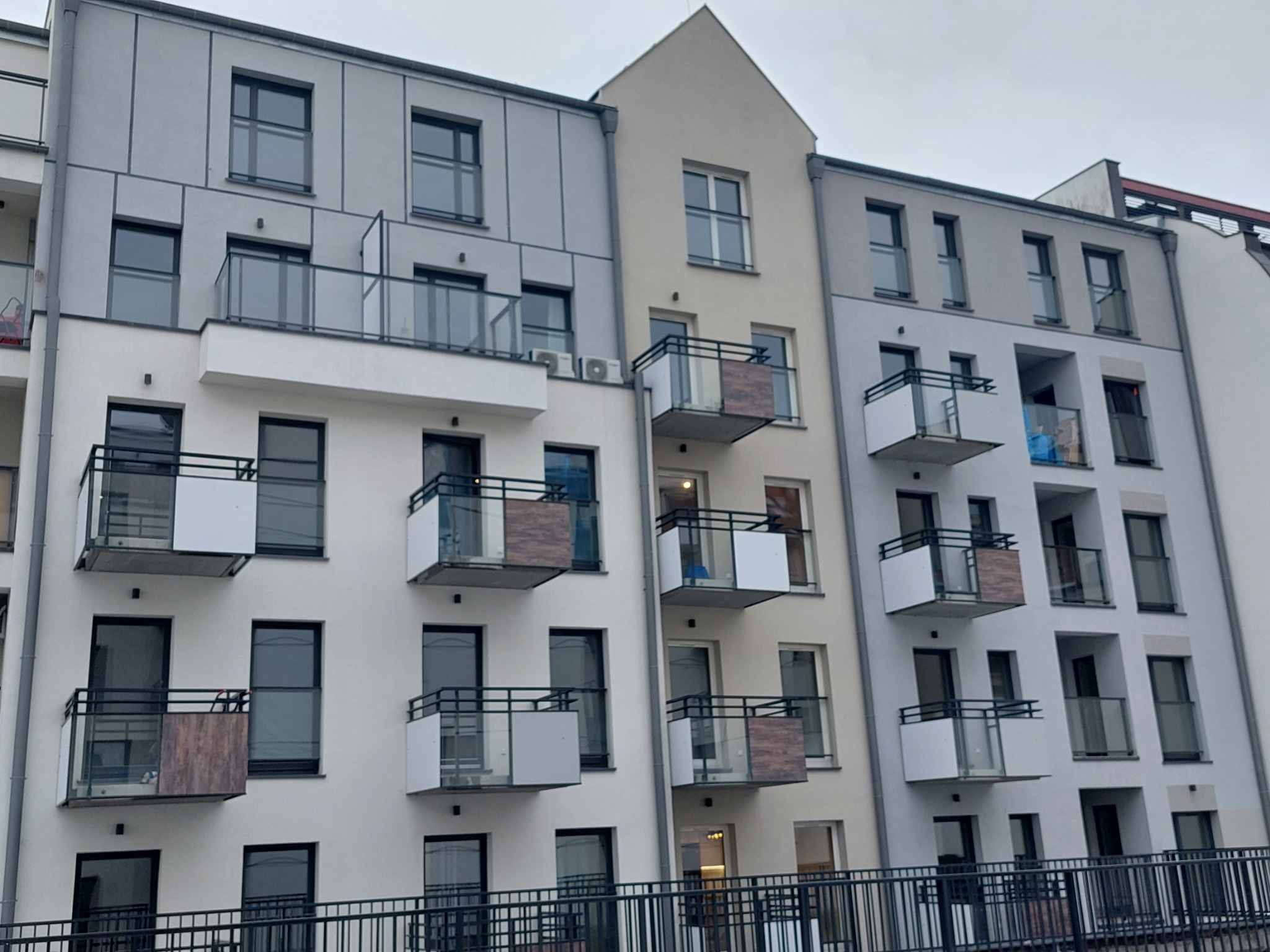 Fasada nowoczesnego budynku wielorodzinnego z balkonami o zróżnicowanym wykończeniu balustrad: szklane, metalowe i z panelami imitującymi drewno, widok z dołu przez metalową barierkę.