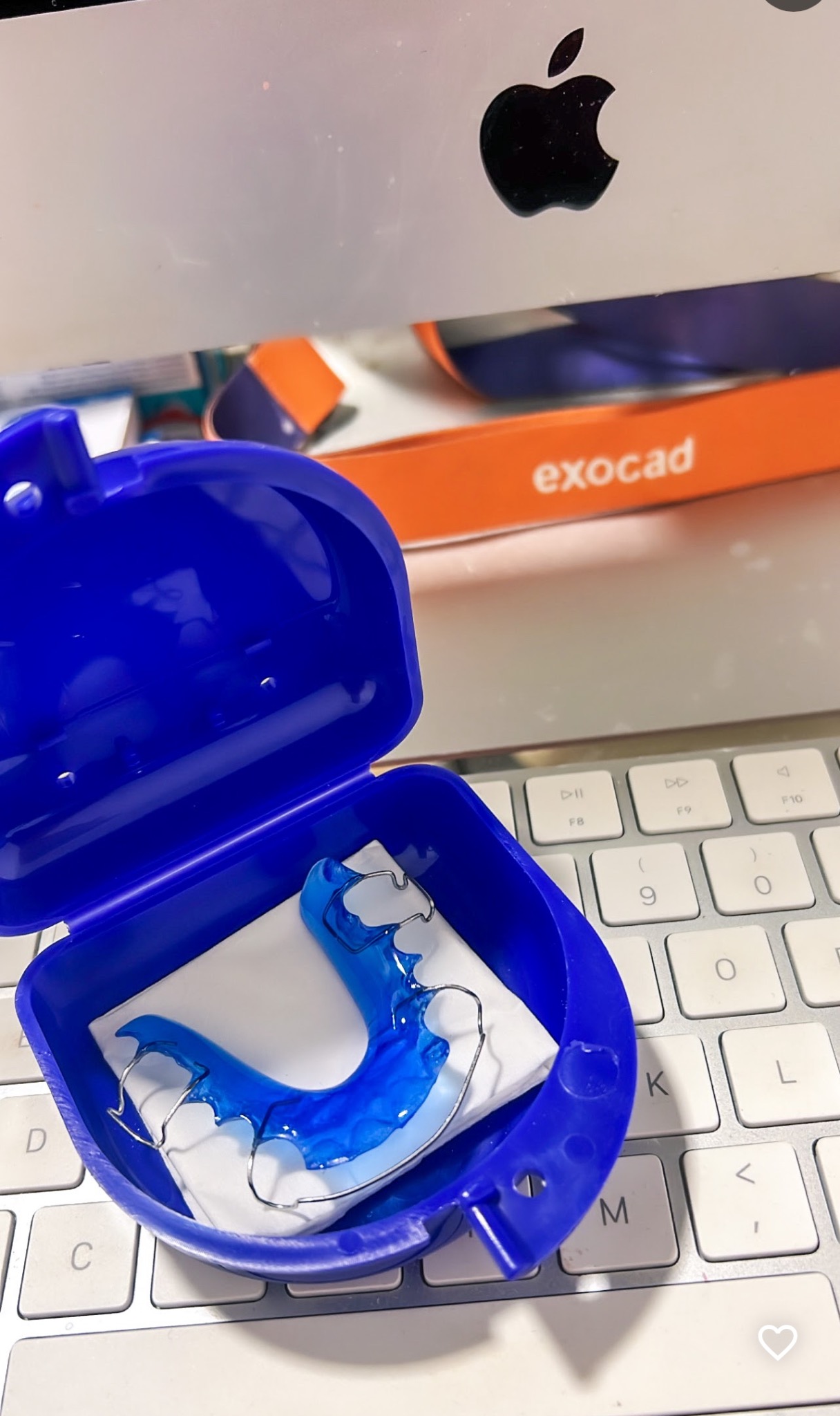 Niebieska, akrylowa szyna relaksacyjna z metalowymi elementami w otwartym, ciemnoniebieskim pudełku, leżąca na białej klawiaturze Apple, z widocznym logo Apple na ekranie komputera w tle...
