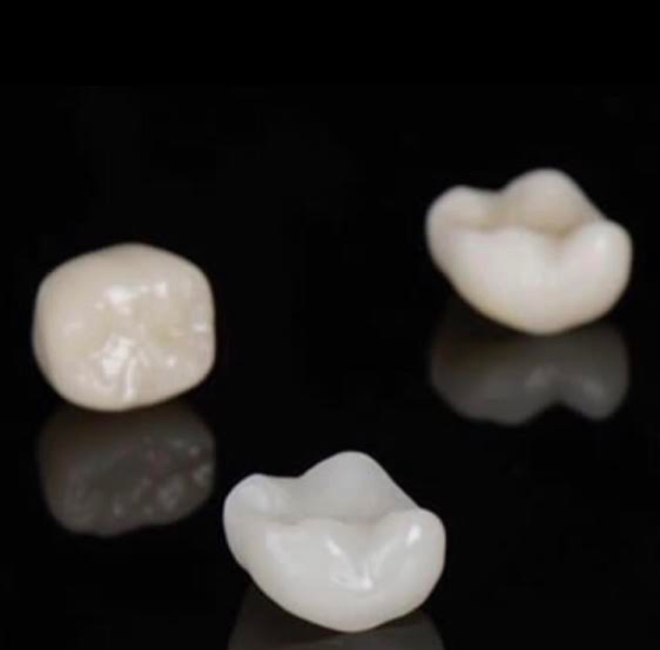 Trzy ceramiczne korony zębowe na czarnym, błyszczącym tle, widoczne odbicia światła.