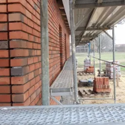 Ceglana ściana budynku w trakcie budowy, widoczna z poziomu rusztowania, z paletami cegieł w tle.