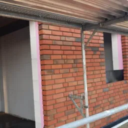 Wznoszenie ścian zewnętrznych z cegły klinkierowej na nowo budowanym domu, widoczne rusztowanie i ocieplenie styropianem.