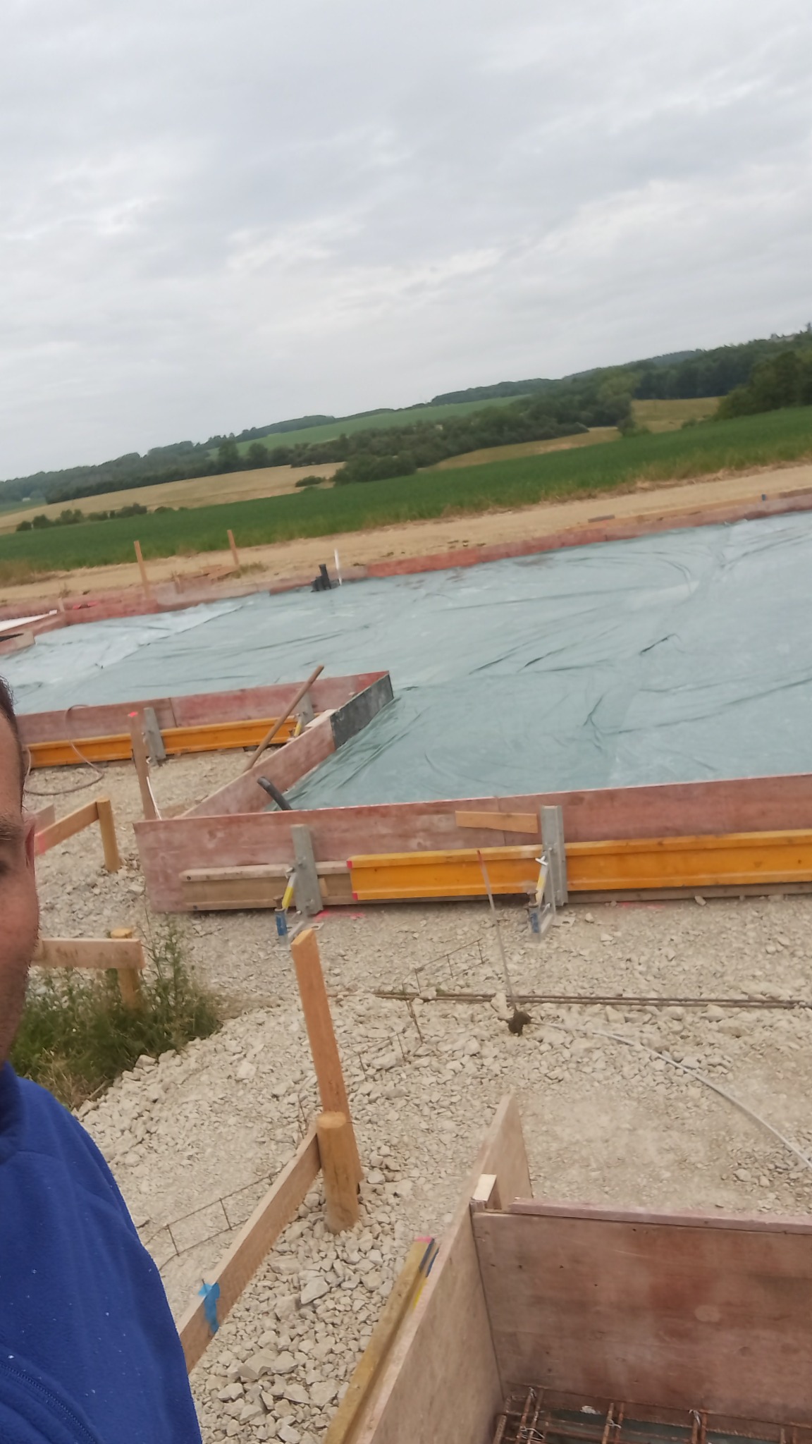 Przygotowanie terenu pod wylewkę fundamentów z drewnianym szalunkiem, zabezpieczone folią budowlaną, widok z perspektywy osoby stojącej obok.