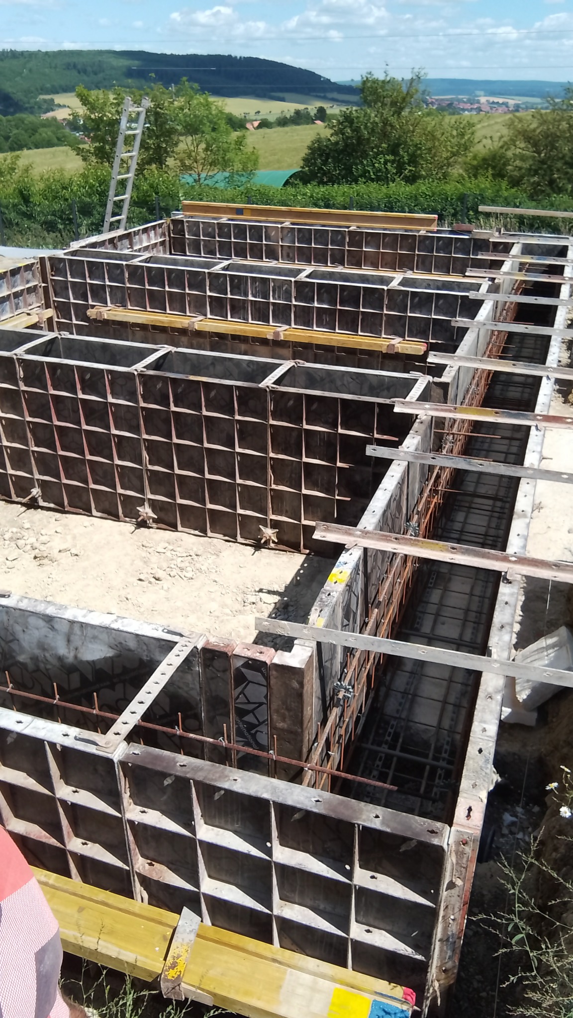 Szczegółowe ujęcie szalunku fundamentów z widocznym zbrojeniem i elementami mocującymi, na tle zieleni i błękitnego nieba.