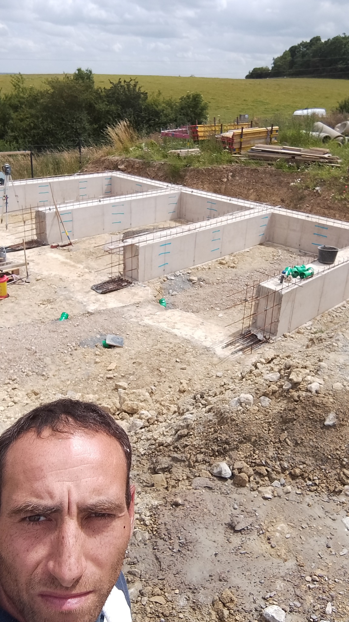 Szare betonowe ściany fundamentowe z wystającą stalą zbrojeniową na placu budowy, z widocznym fragmentem twarzy budowlańca w dolnej części kadru.