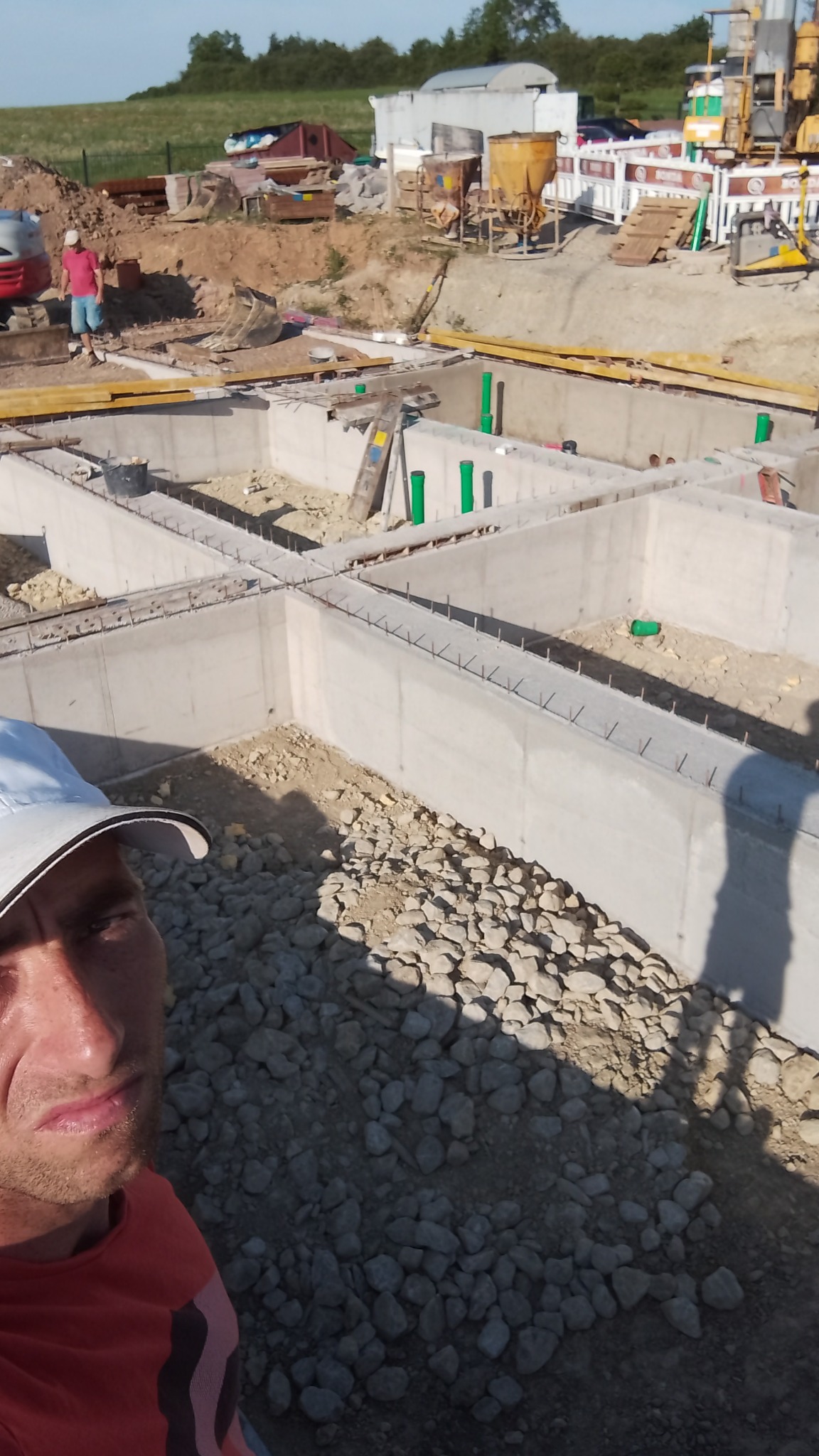 Wykop pod fundamenty domu z widocznymi betonowymi ścianami szalunkowymi, zbrojeniami i instalacją kanalizacyjną, z perspektywy selfie pracownika w kasku, z maszynami budowlanymi w tle.