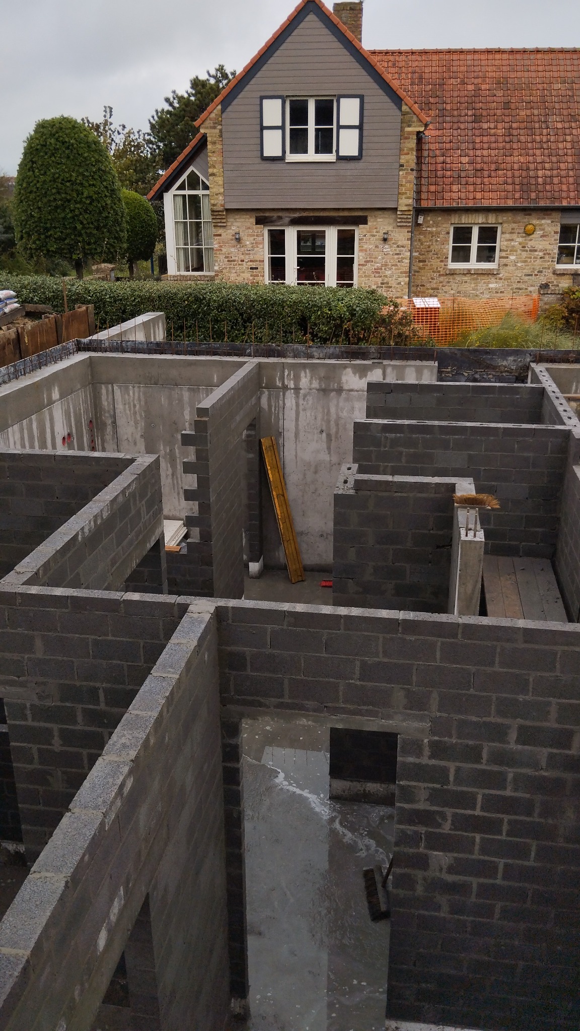 Wykop fundamentowy z częściowo wzniesionymi ścianami z bloczków betonowych, widoczna drewniana łata murarska oparta o ścianę, w tle dom jednorodzinny w stylu angielskim.