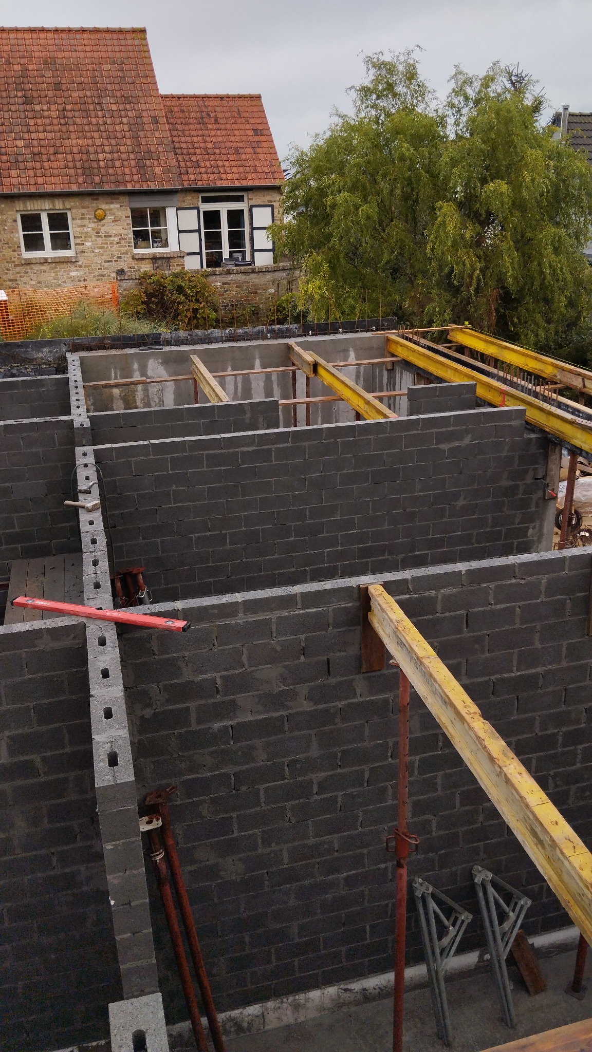 Wznoszenie ścian fundamentowych z betonowych bloczków z użyciem żółtych belek wsporczych i poziomicy w kolorze czerwonym, widok z góry na plac budowy z domem w tle.