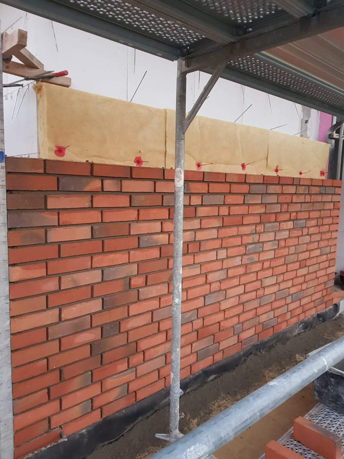 Ściana z czerwonej cegły w trakcie wznoszenia, widoczne ocieplenie budynku i rusztowanie.
