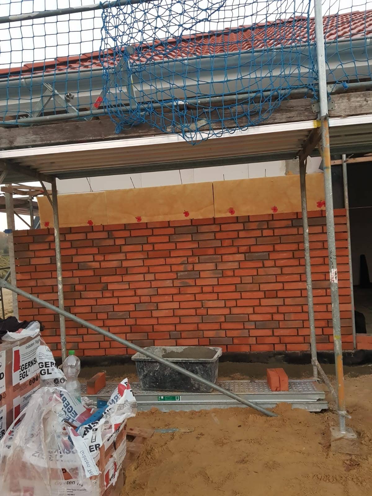 Świeżo wymurowana ściana z cegły klinkierowej na budowie domu, widoczna z rusztowania, z zapasem cegieł i pojemnikiem z zaprawą murarską w pierwszym planie.