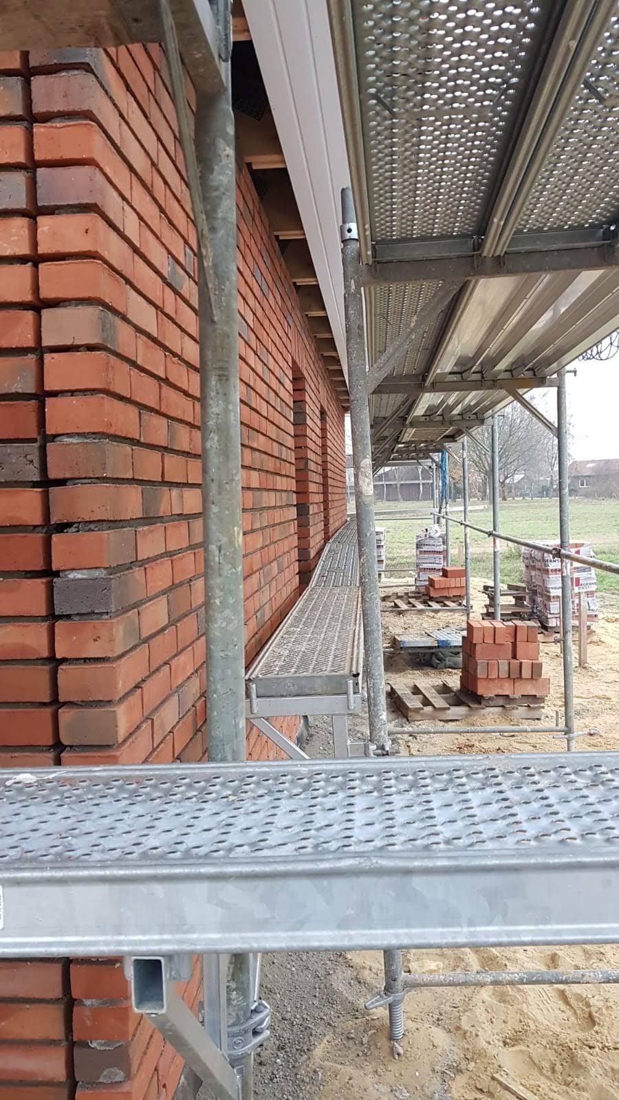 Ceglana ściana budynku w trakcie budowy, widoczna z poziomu rusztowania, z paletami cegieł w tle.