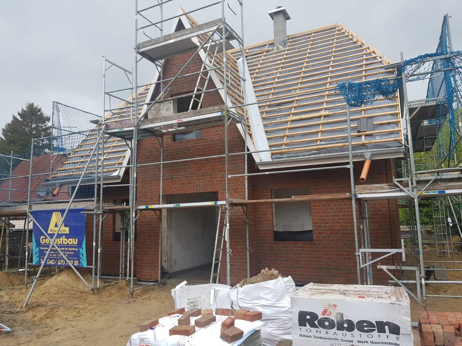 Budowa domu jednorodzinnego w Jaworze, widoczne rusztowania, częściowo wymurowane ściany z czerwonej cegły, przygotowana konstrukcja dachu z deskowaniem i papą, oraz materiały budowlane na placu...