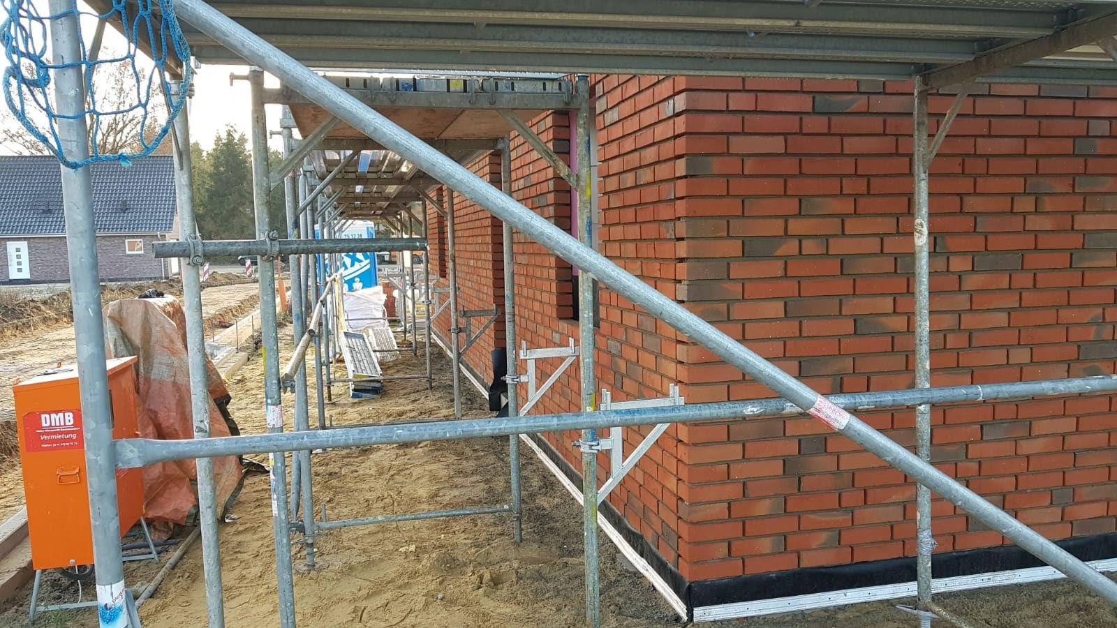 Wznoszenie ściany zewnętrznej z czerwonej cegły na placu budowy, widoczne rusztowanie, pomarańczowa skrzynka narzędziowa DMB Vermietung i częściowo zasłonięty fundament.