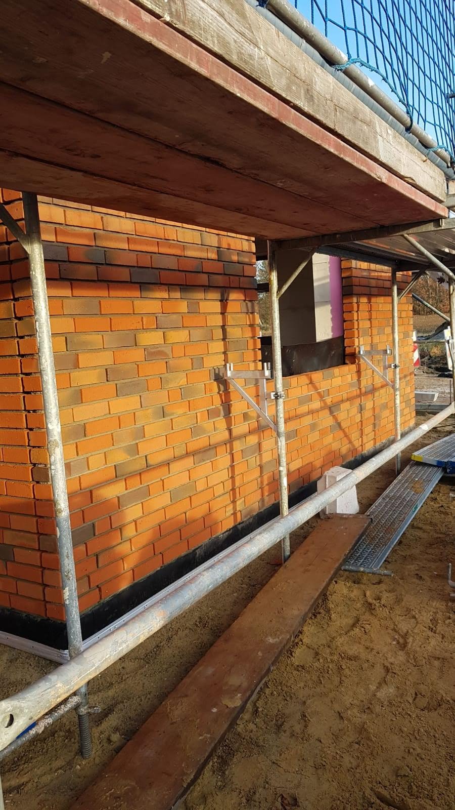 Świeżo wymurowana ściana z cegły klinkierowej w odcieniach pomarańczy i brązu, widoczna spod rusztowania. Fragment okna z izolacją z różowego styropianu. Zabezpieczająca siatka na górze.