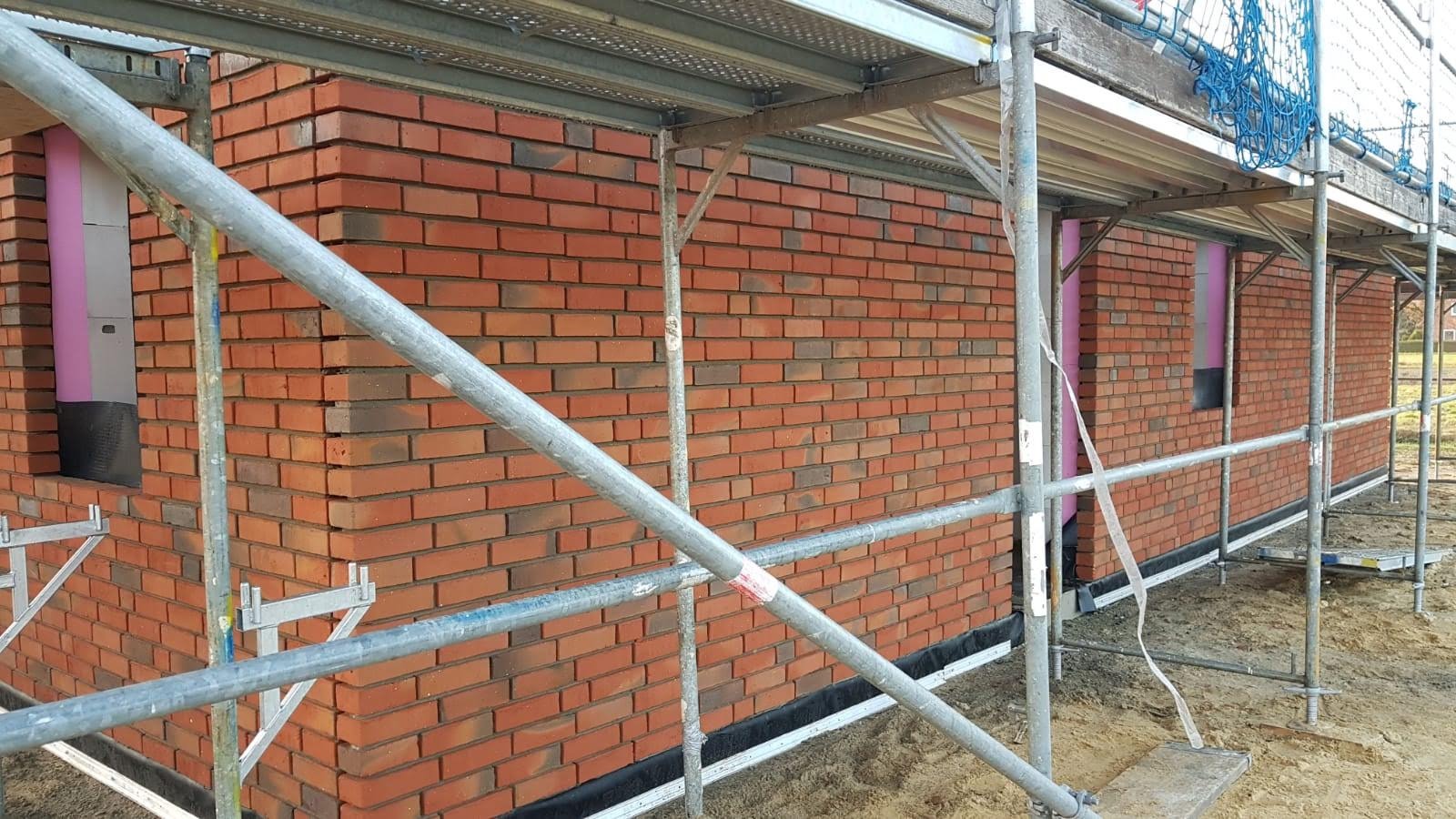 Wznoszenie ściany z czerwonej cegły na budowie domu, widoczne rusztowanie i ocieplenie wokół okien.