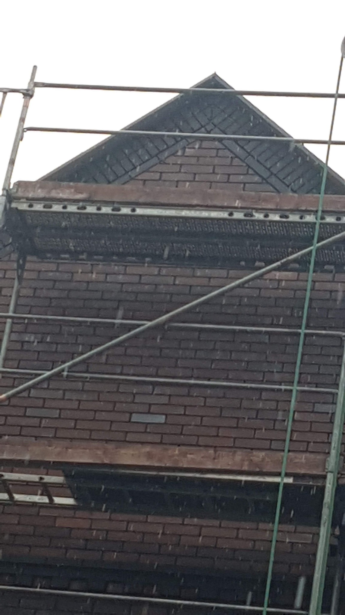 Fragment murowanej ściany budynku z ciemnej cegły, widoczny z poziomu rusztowania, z fragmentem dachu pokrytego ciemną dachówką.