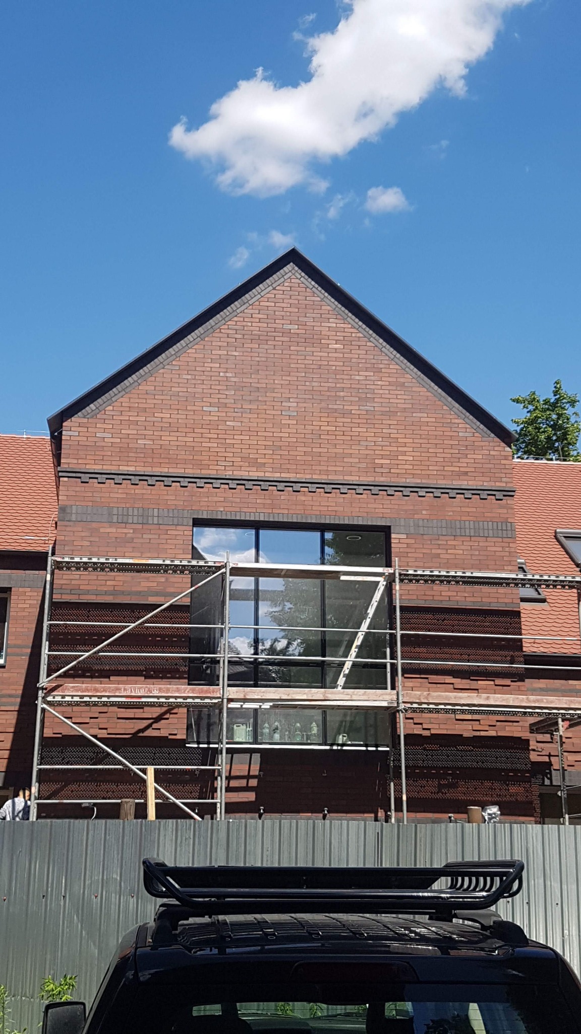 Wznoszona ściana budynku z czerwonej cegły z dużym oknem, widoczne rusztowanie i fragment samochodu na pierwszym planie.