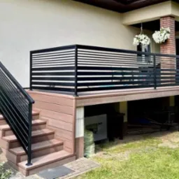 Nowoczesna balustrada balkonowa z poziomych profili, czarna, zamontowana na drewnianym podeście z schodami, widok na dom z wiszącymi kwiatami.