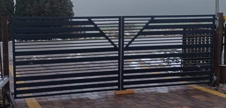 Nowoczesna, dwuskrzydłowa brama wjazdowa z poziomymi profilami, pomalowana na czarno, z geometrycznym wzorem trójkąta w górnej części, zamontowana na posesji z brukowaną nawierzchnią.