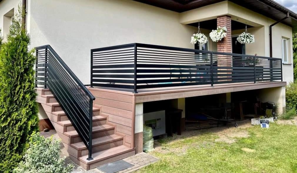 Nowoczesna balustrada balkonowa z poziomych profili, czarna, zamontowana na drewnianym podeście z schodami, widok na dom z wiszącymi kwiatami.