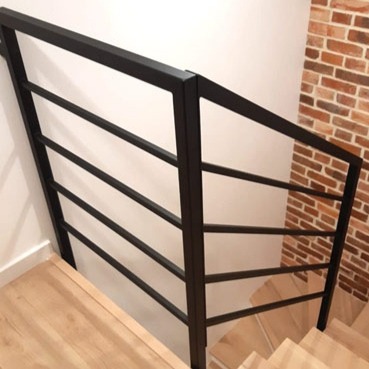 Nowoczesna, czarna, metalowa balustrada schodowa z poziomymi prętami, zamontowana na drewnianych schodach, z fragmentem ściany z cegły w tle.