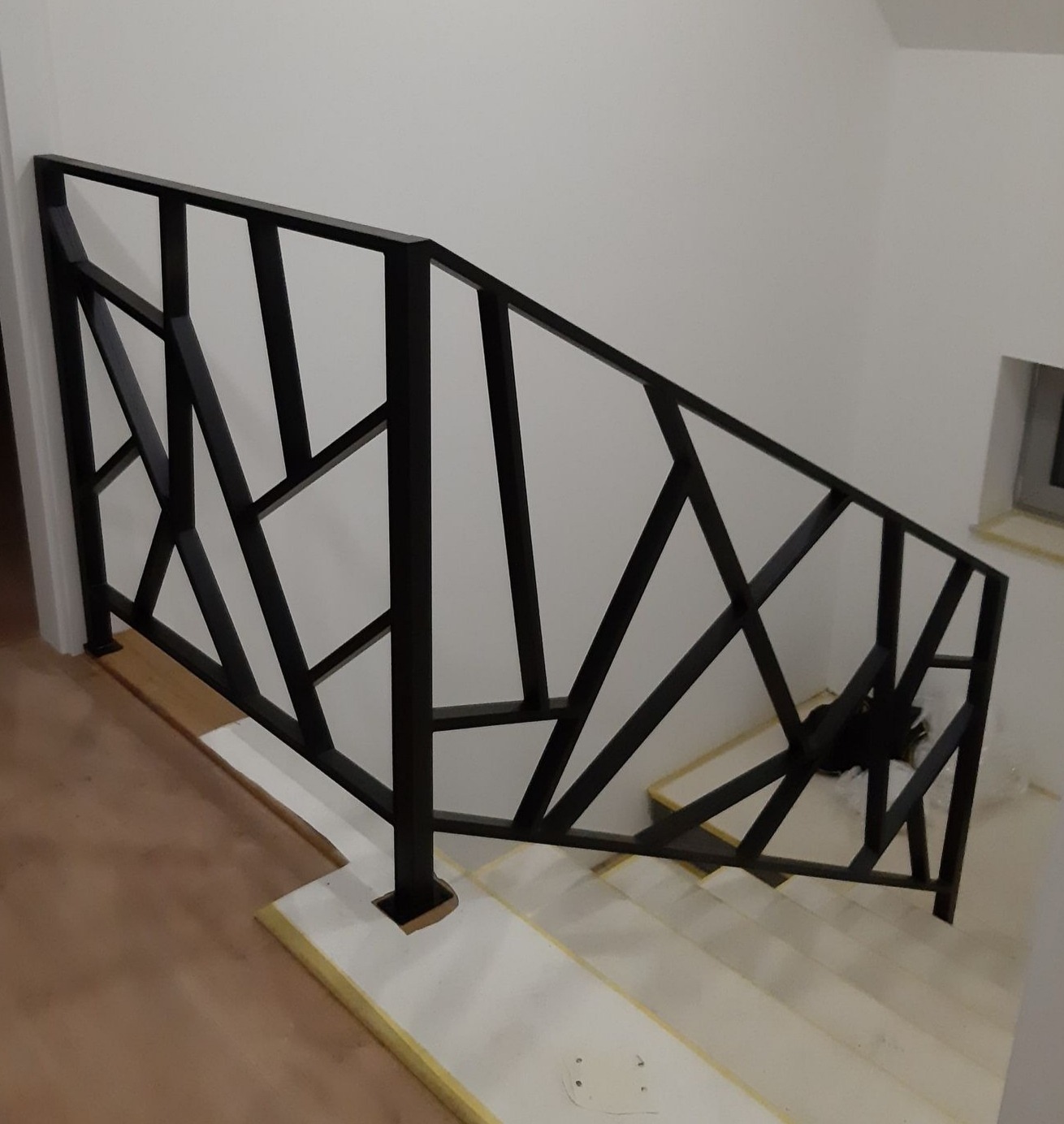 Czarna, metalowa balustrada schodowa o geometrycznym wzorze, widok z góry na schody w trakcie wykończenia, białe ściany w tle.