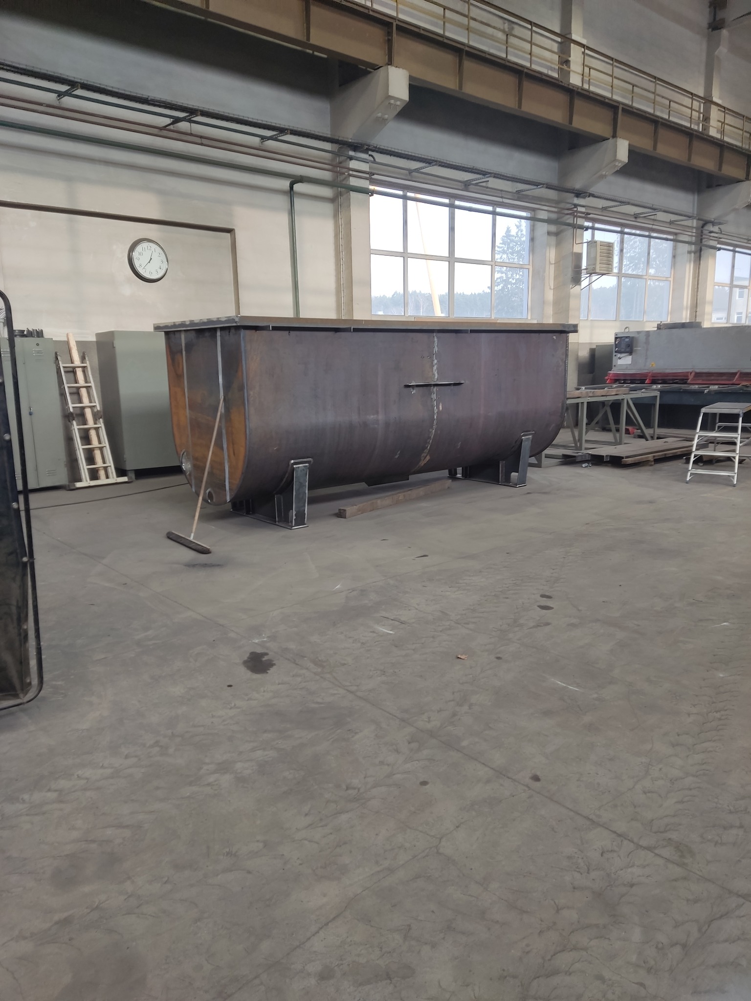 Duży, metalowy zbiornik o zaokrąglonym kształcie, na niskich nóżkach, stoi w hali produkcyjnej. Obok zbiornika oparta o niego zmiotka. W tle widoczne okna i inne urządzenia.