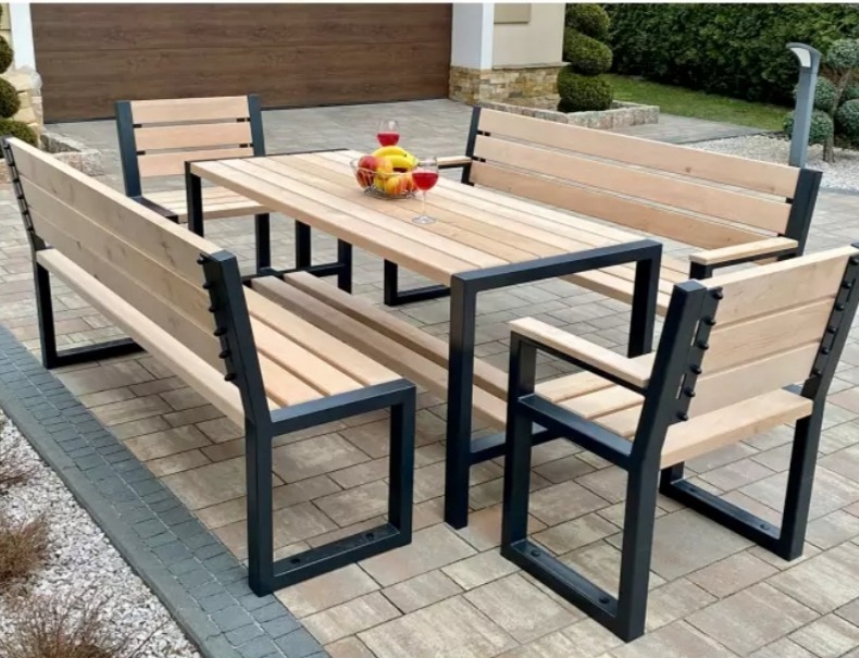 Zestaw mebli ogrodowych: stół z blatem z jasnego drewna i cztery krzesła/ławki z metalowymi, grafitowymi ramami, na stole misa z owocami i dwa kieliszki, tło: kostka brukowa i budynek.