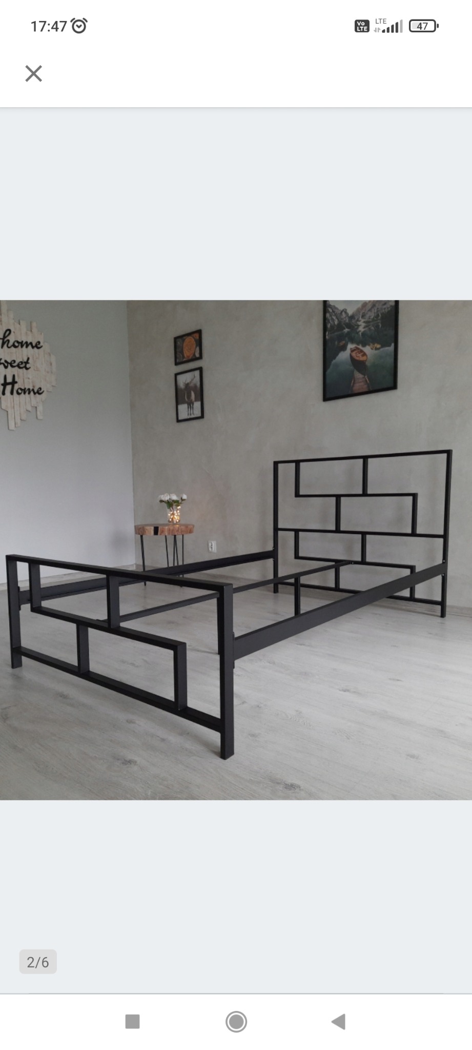 Metalowa rama łóżka z geometrycznym zagłówkiem, czarna, na tle szarej ściany z dekoracjami i jasną podłogą imitującą drewno.